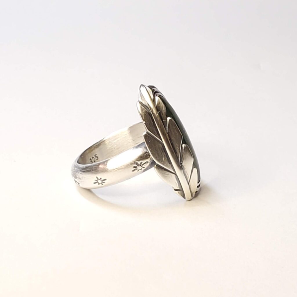 A view of the sided of this ring, from the right. A sterling silver ring set with an oval, green jade. Hugging the left side of the stone is a formed silver feather made in the Southwest style.