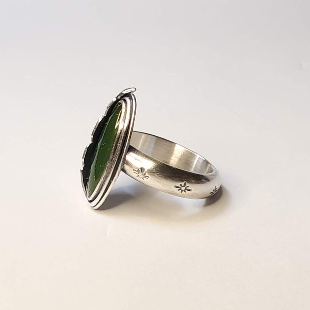 A view of the side of this ring from the left. A sterling silver ring set with an oval, green jade. Hugging the left side of the stone is a formed silver feather made in the Southwest style.