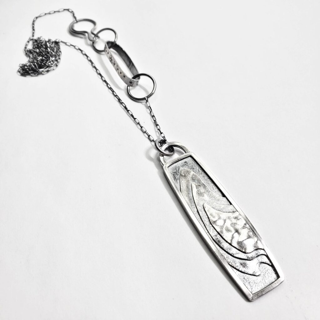 A closeup of one of the terrain pendants. This one is longer than it is wide, with multiple layers of silver stacked and fused to each other to create that appears to be a stylized contour map.