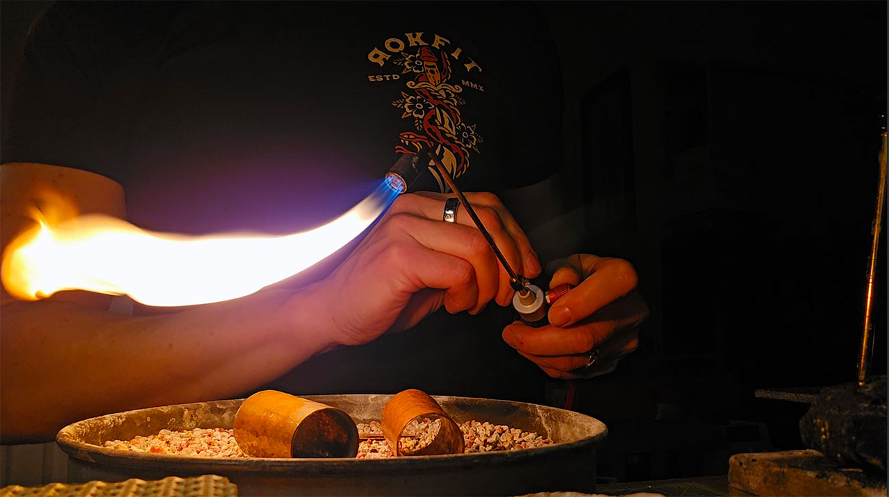 The artist's hands holding a lit torch, preparing to anneal some metal.
