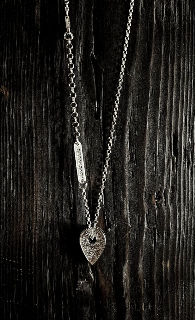 A necklace featuring a map marker planchette pendant made with reticulated silver and framed in a narrow sterling silver strip. A silver tag with the word, "Adventure" is shown further up the chain.